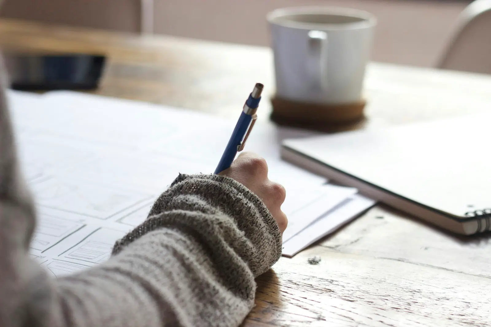 A hand in a knit sweater filling out paperwork with a pen at a wooden table, a coffee mug nearby
